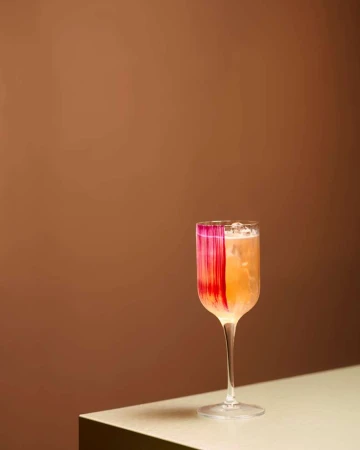 A tall glass cocktail with ice and a red painted brushstroke on the rim, set on a light table against a warm brown wall.