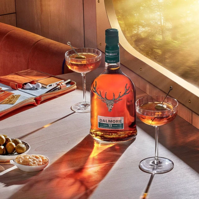 Bottle of Dalmore whisky with two stemmed cocktail glasses, a leather case, and a dish of limes on a bright wooden table.
