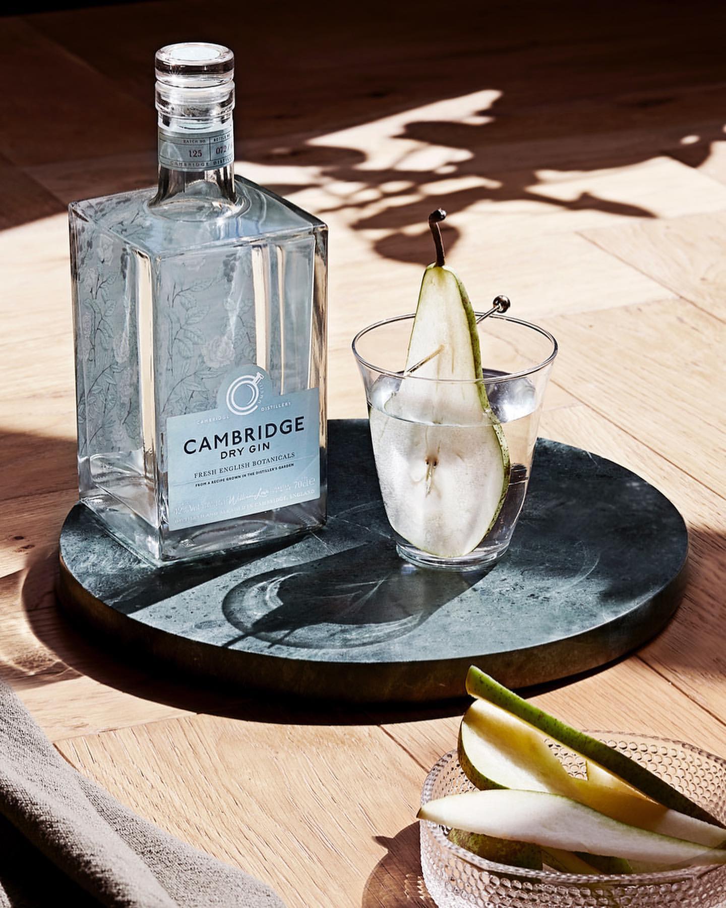 Still life with a gin bottle and a cocktail glass garnished with a pear slice, set on a round marble tray on a wooden table.