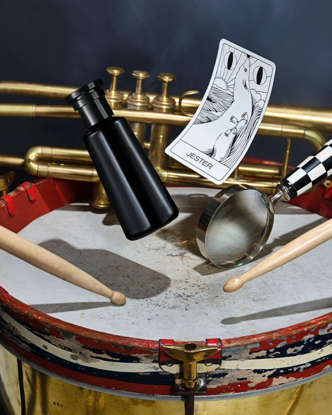Still life photograph features a black bottle, playing card titled Jester, and drumsticks on a drum with a brass trumpet background.