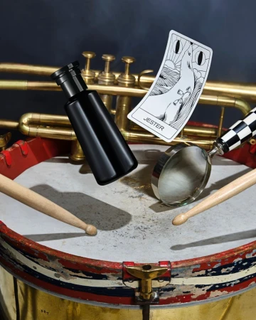 Still life photograph features a black bottle, playing card titled Jester, and drumsticks on a drum with a brass trumpet background.