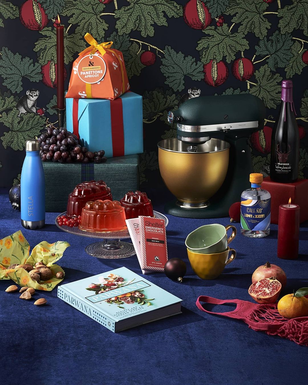 Still life photography featuring a table set with gifts, a cookbook, wine, baking equipment, and food, against a patterned backdrop.