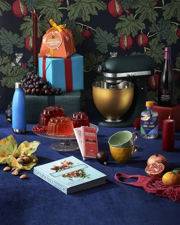 Still life photography featuring a table set with gifts, a cookbook, wine, baking equipment, and food, against a patterned backdrop.