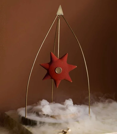 Red star-shaped object on a gold metal frame, centered over a white smoky base against a gradient brown background, creating an artistic, unique display.