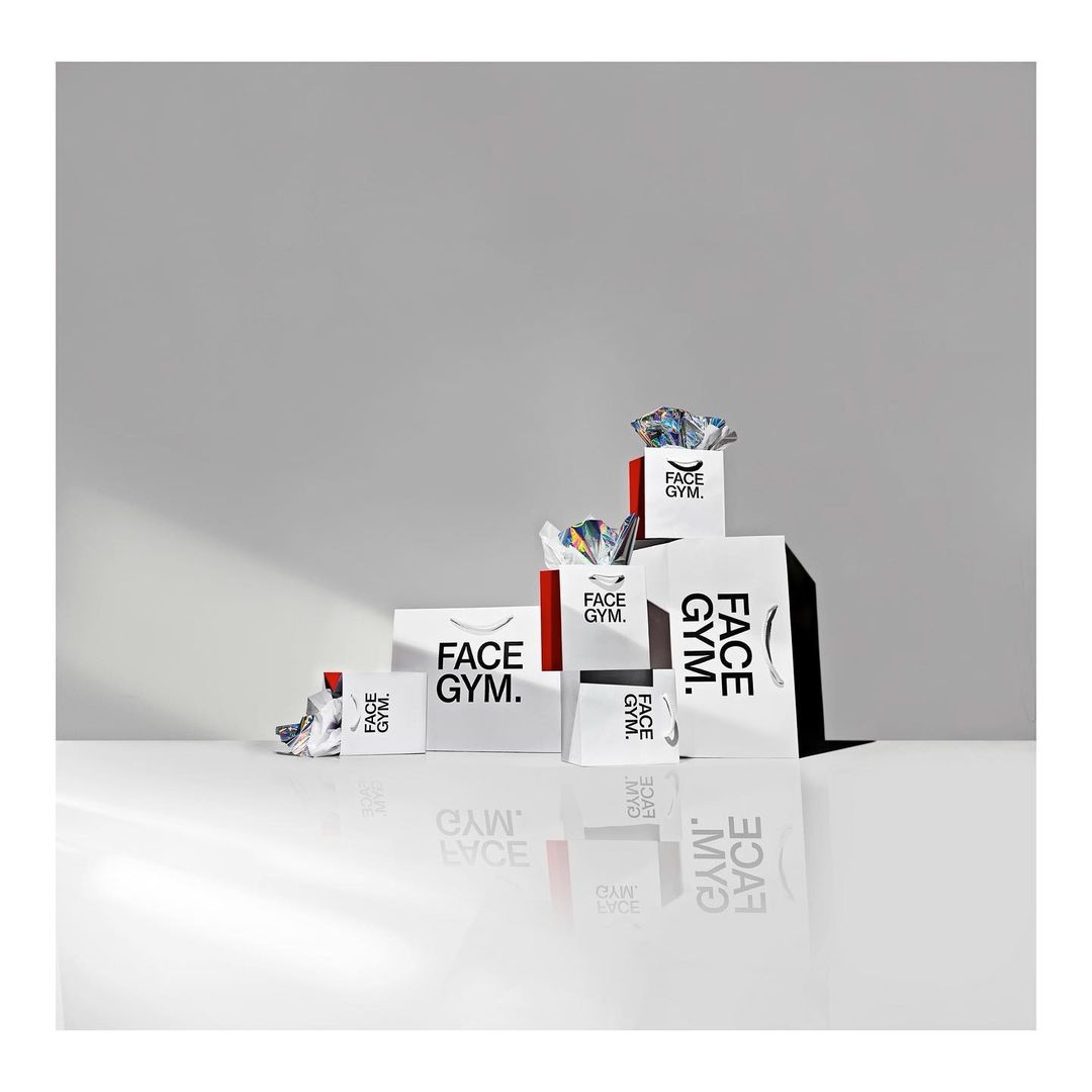 Various sizes of Face Gym branded shopping bags arranged on a white surface against a graduated gray background in studio lighting.