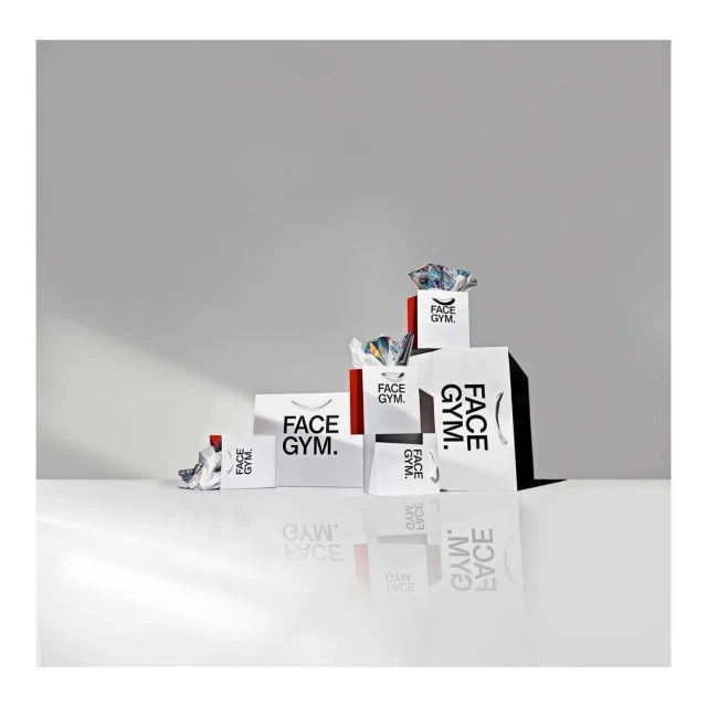 Various sizes of Face Gym branded shopping bags arranged on a white surface against a graduated gray background in studio lighting.