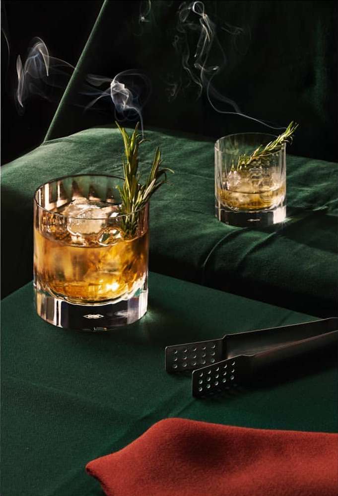 Two whiskey cocktails with ice and rosemary sprigs on a dark green velvet surface with a cocktail strainer, creating a sophisticated ambiance.