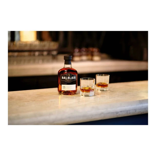 Bottle of Balblair scotch whisky with two glasses of whisky, on a counter in a dimly lit bar setting with glasses in background.