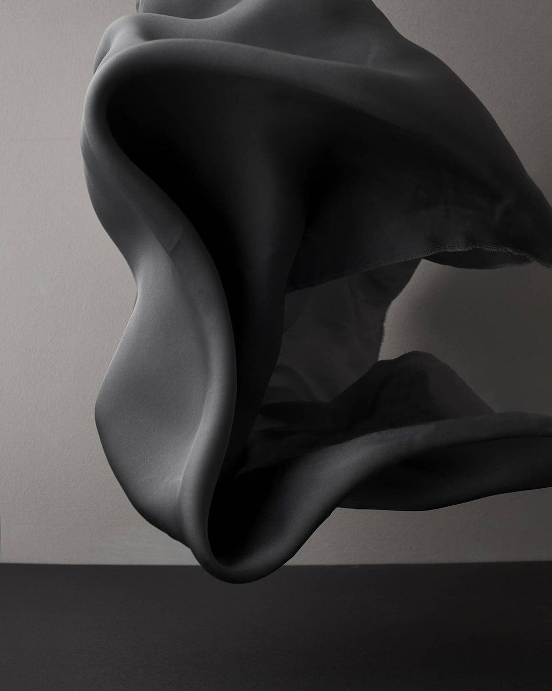 Abstract monochrome photo of flowing draped fabric with smooth, fluid forms and soft shadows against a minimal gray background.