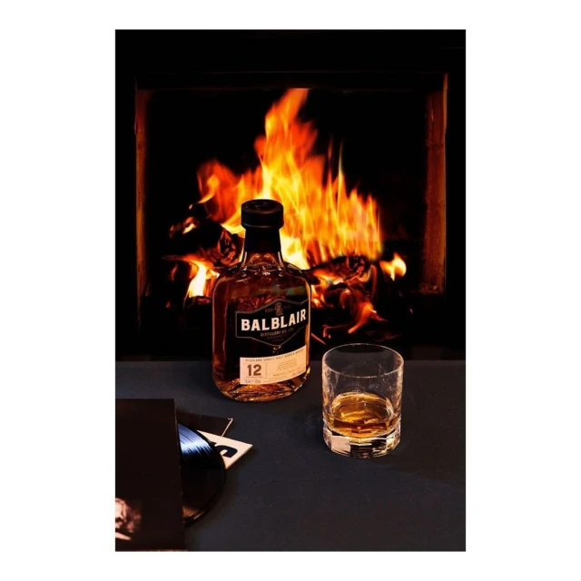 A bottle of Balblair whisky and a glass with a vinyl record in front of a fireplace with burning logs, creating a cozy scene.
