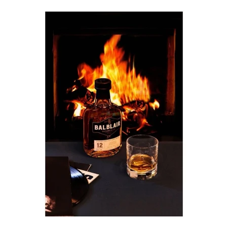 A bottle of Balblair whisky and a glass with a vinyl record in front of a fireplace with burning logs, creating a cozy scene.