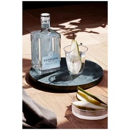 A clear glass bottle of Cambridge Dry Gin and a cocktail garnished with a pear slice on a dark round tray.