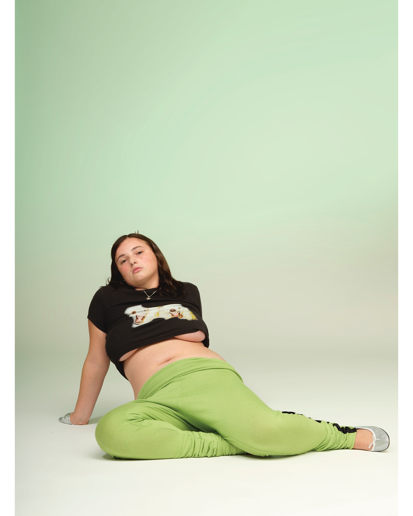Young adult woman poses in a photo studio wearing a graphic t-shirt and green sweatpants. The image has a soft, light-green background.