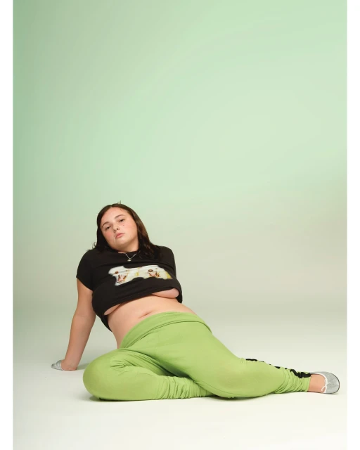 Young adult woman poses in a photo studio wearing a graphic t-shirt and green sweatpants. The image has a soft, light-green background.