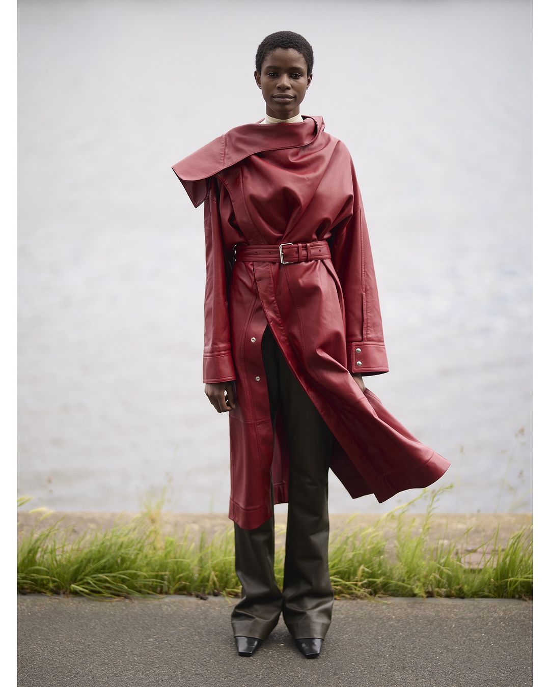 A dark-skinned adult fashion model poses outdoors, wearing a belted long red leather coat, long dark pants, and black shoes.
