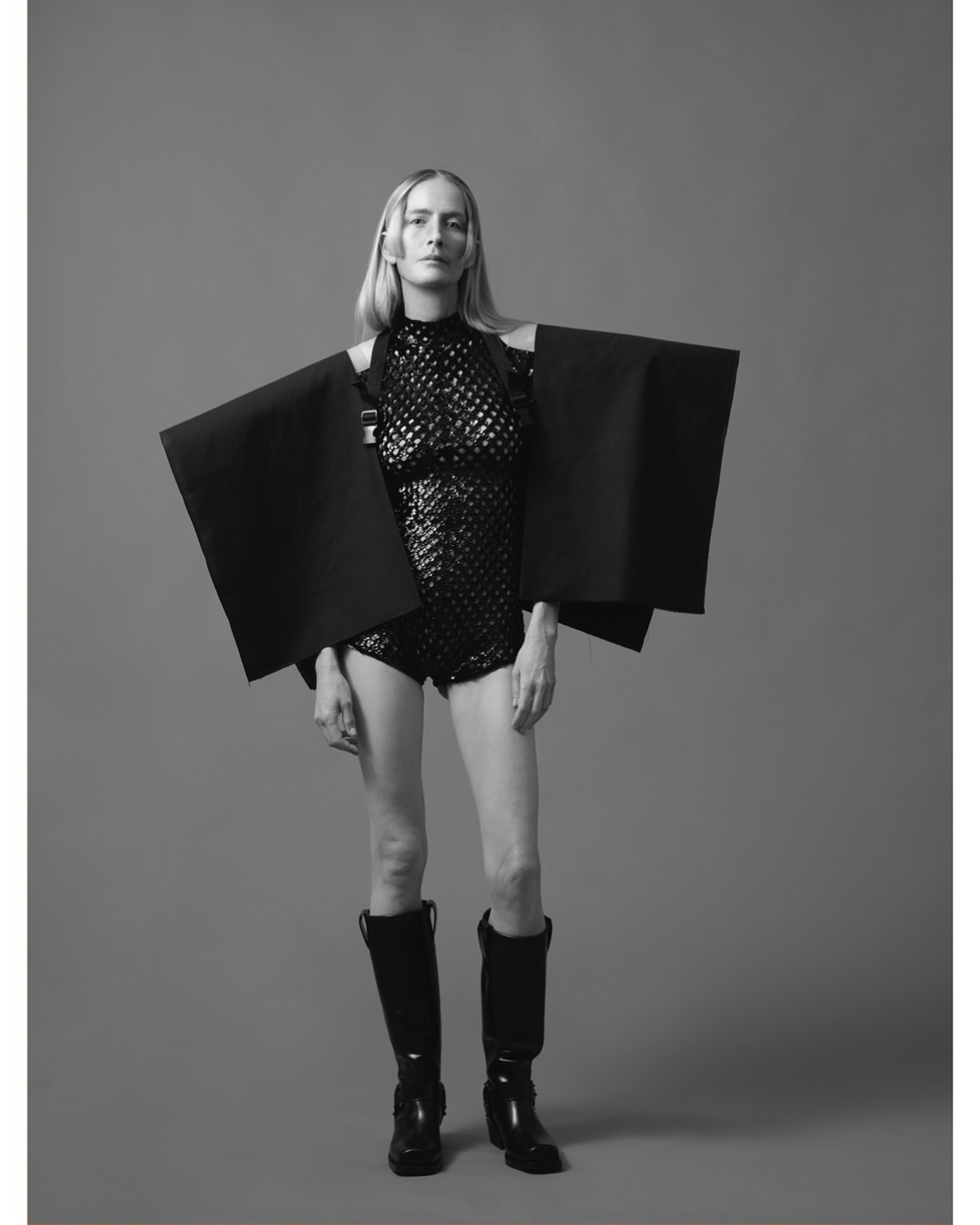Monochrome fashion shot of a woman in a sequined bodysuit with geometric shoulder panels and knee-high boots against a gray background.