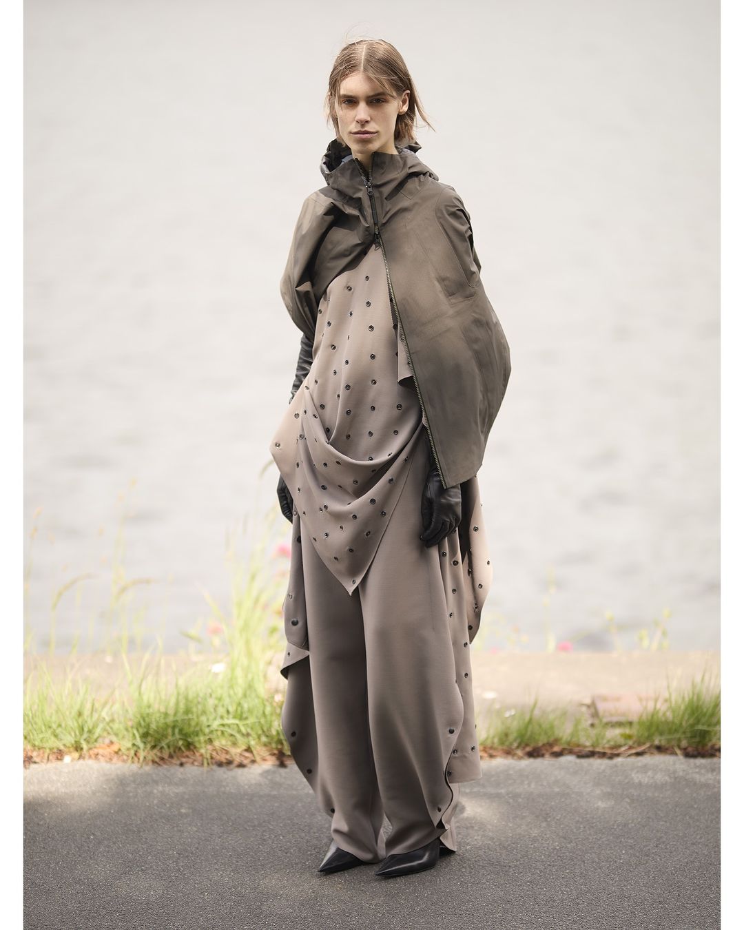A person wearing a neutral tone hooded cape with pants, standing in an outdoor setting near a body of water, for a fashion editorial.