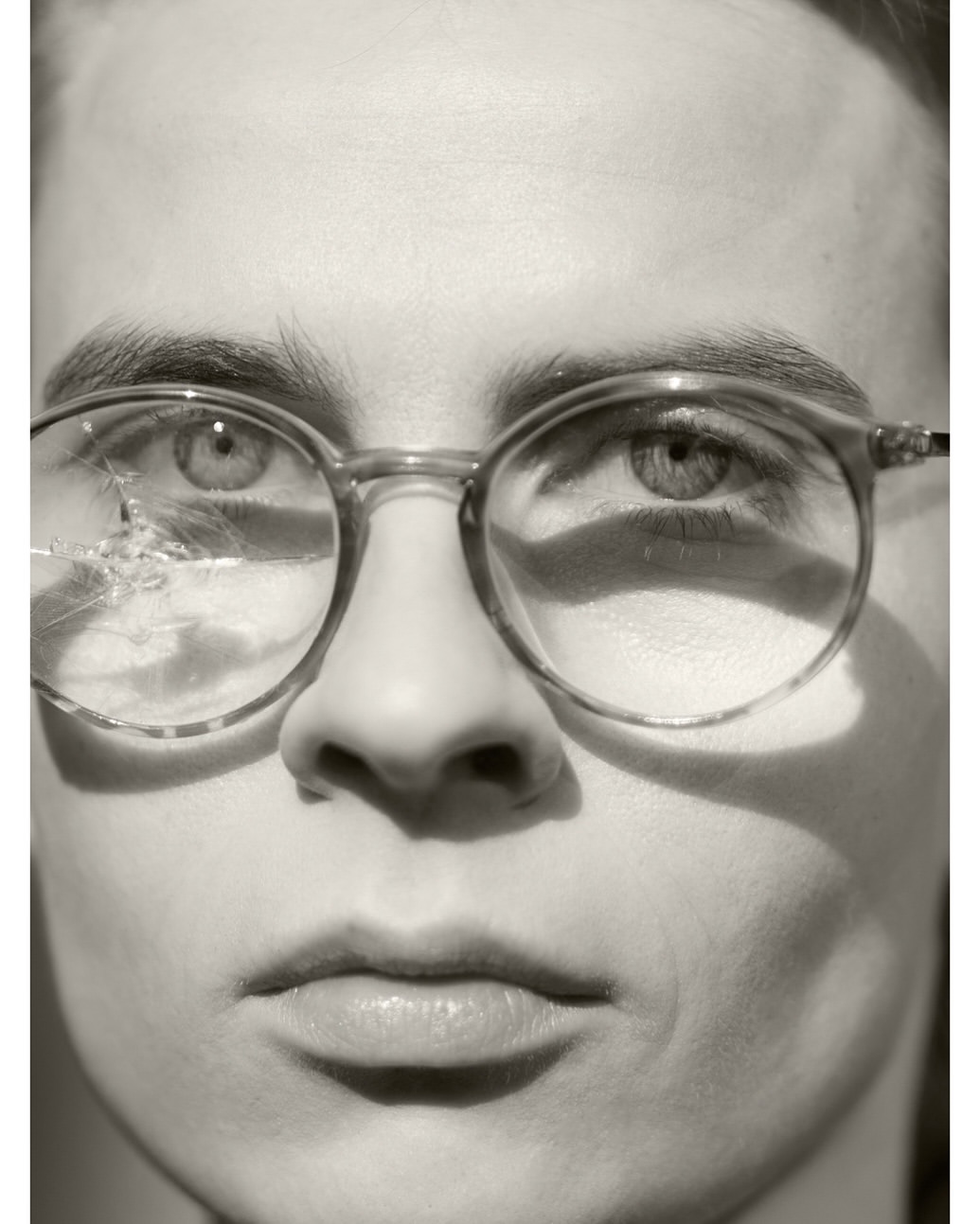 Close-up portrait of a young adult wearing round eyeglasses with a cracked lens, neutral facial expression, black and white image.