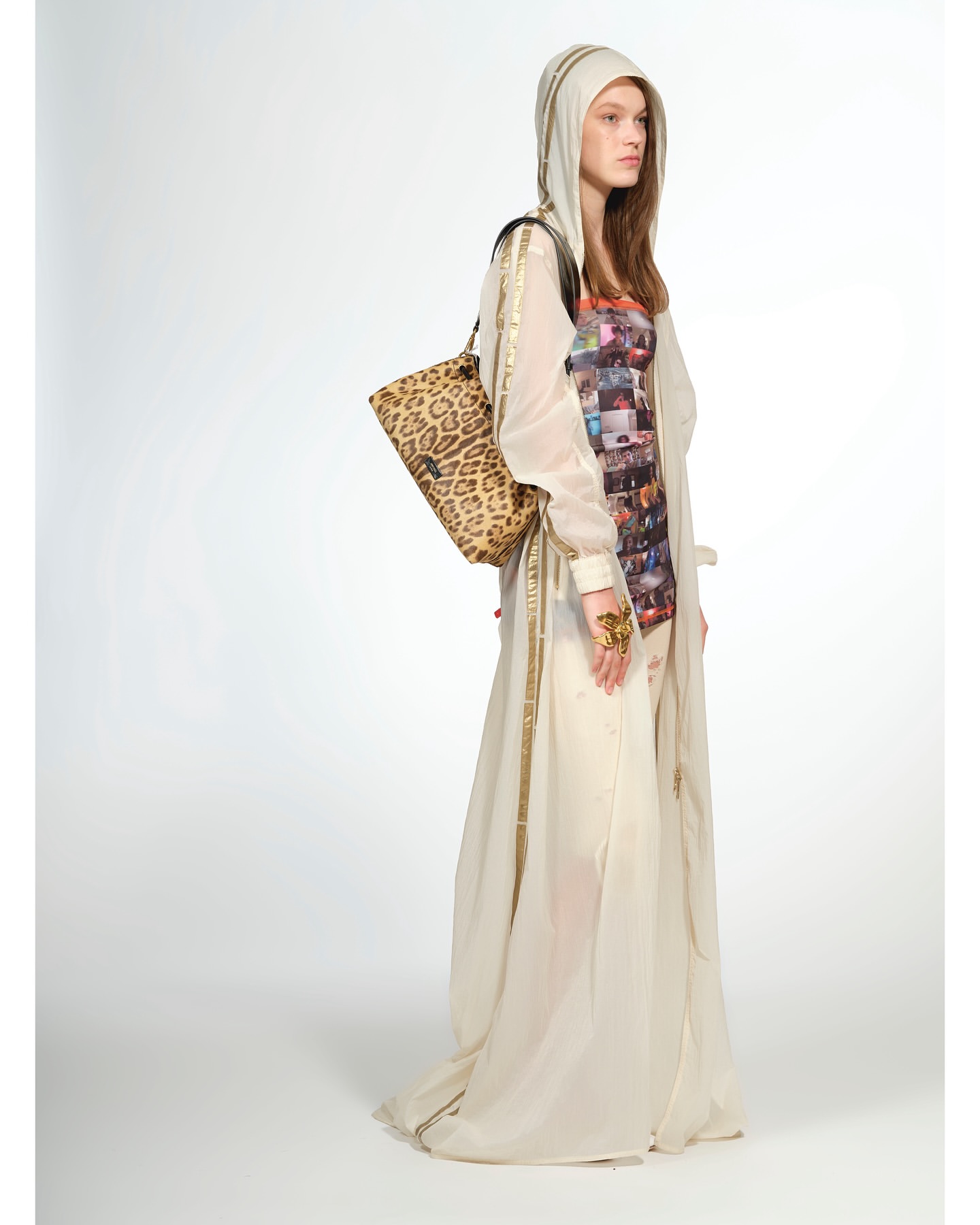 Fashion model in a sheer hooded gown and leopard-print tote bag, posing in a studio with a bright white backdrop. Full-length shot.