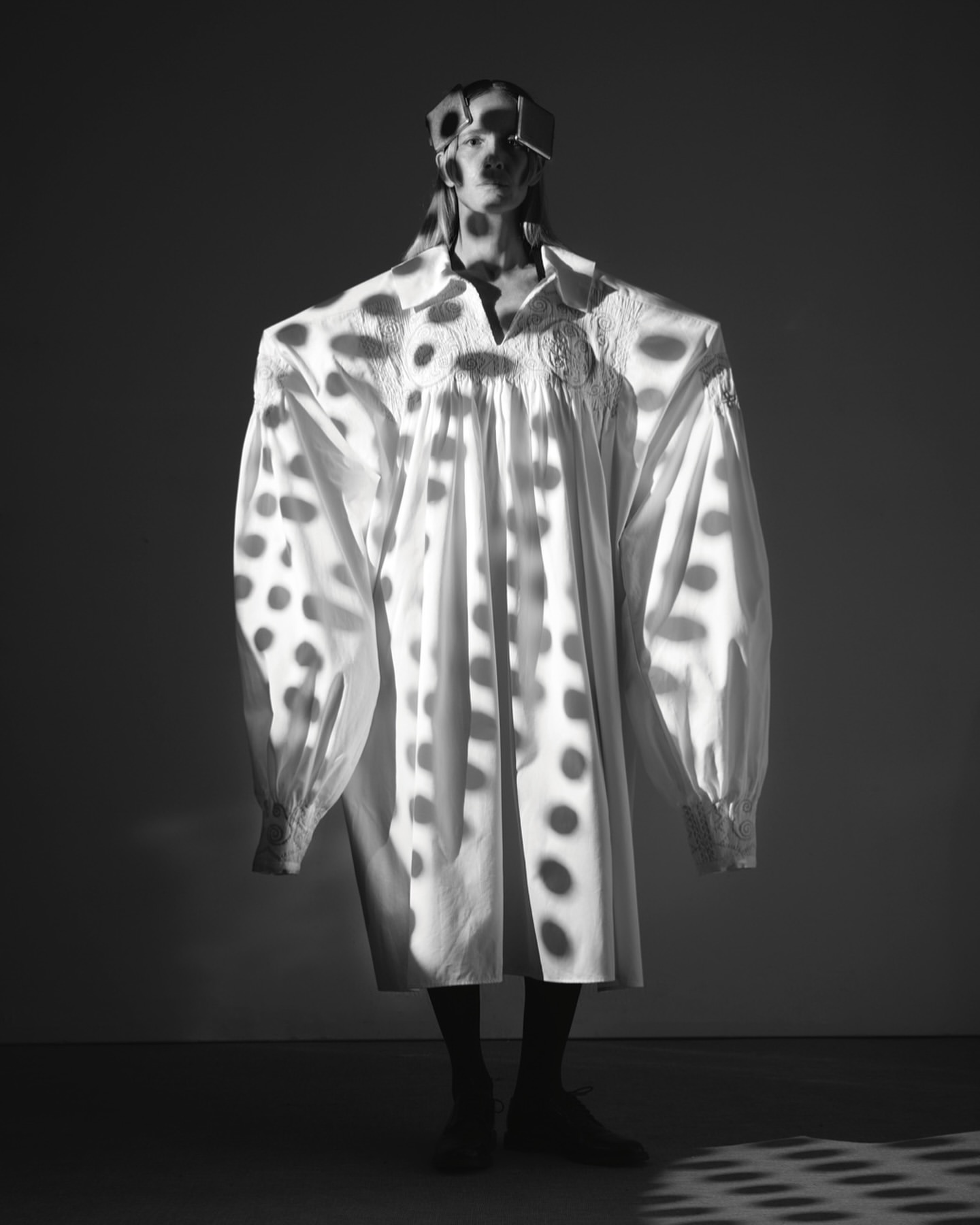 Monochrome portrait of a person wearing an oversized shirt and a box on their head, with polka dot shadows and dark backdrop.