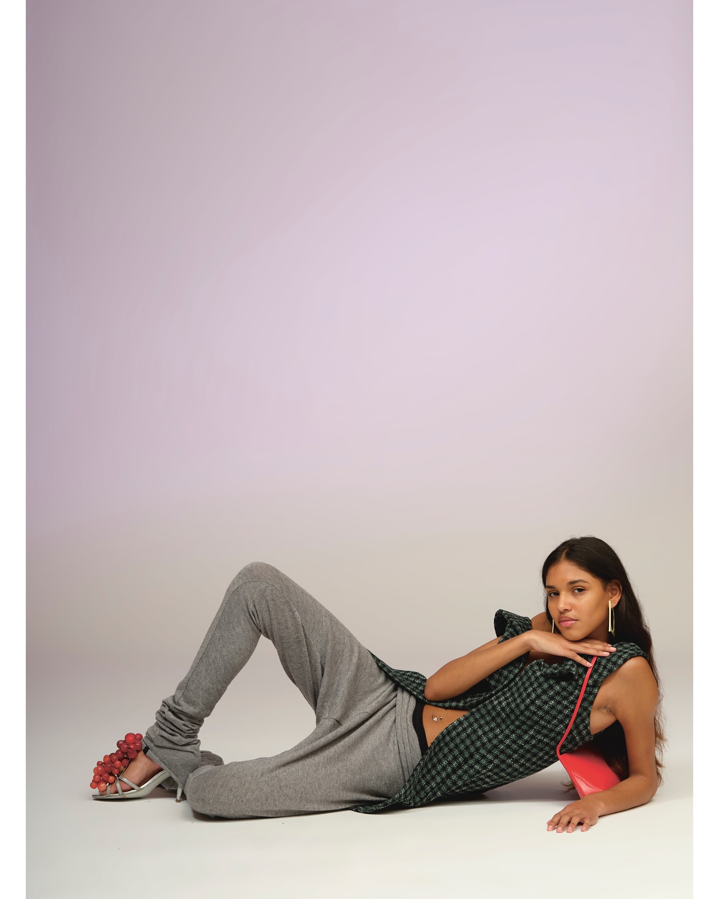 A woman lies on her side wearing grey sweatpants, a plaid sleeveless top, and high heels adorned with red grapes. She poses against a gradient lilac background.