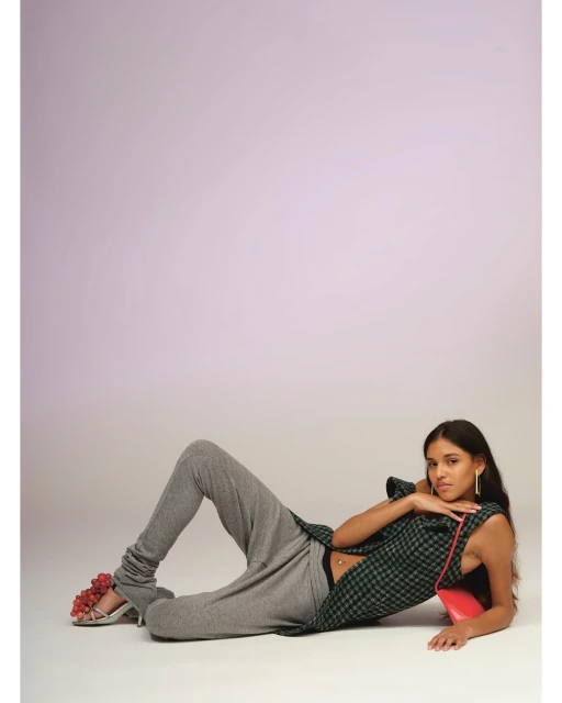 A woman lies on her side wearing grey sweatpants, a plaid sleeveless top, and high heels adorned with red grapes. She poses against a gradient lilac background.