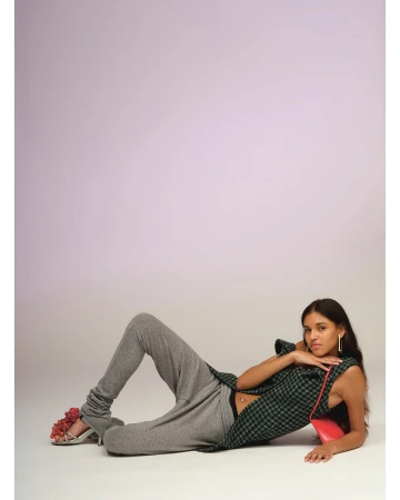 A woman lies on her side wearing grey sweatpants, a plaid sleeveless top, and high heels adorned with red grapes. She poses against a gradient lilac background.