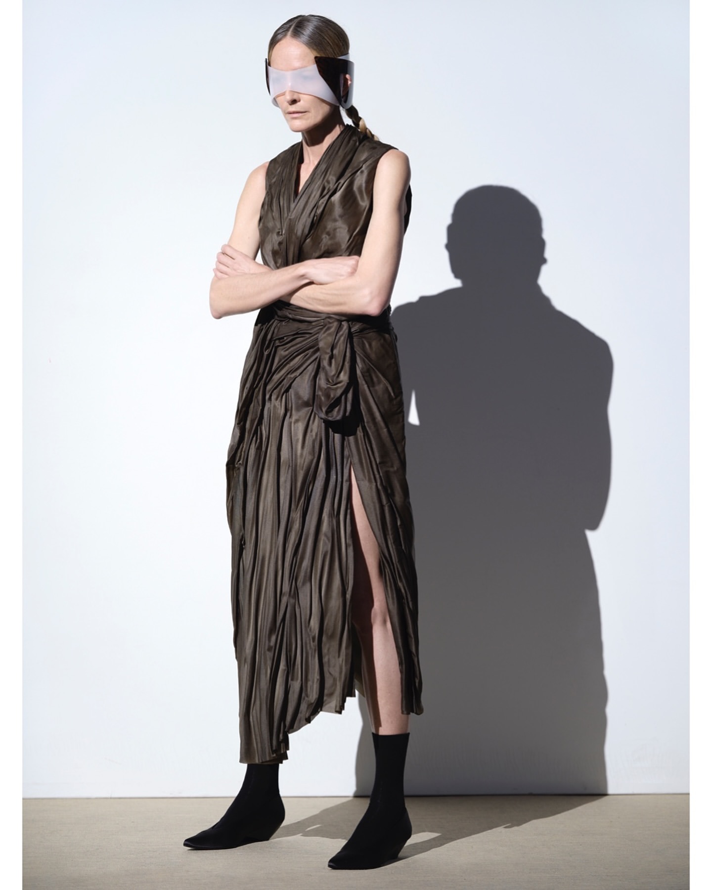 Woman in draped dress and futuristic visor poses against a white background. Fashion shot in a studio with cast shadow detail.