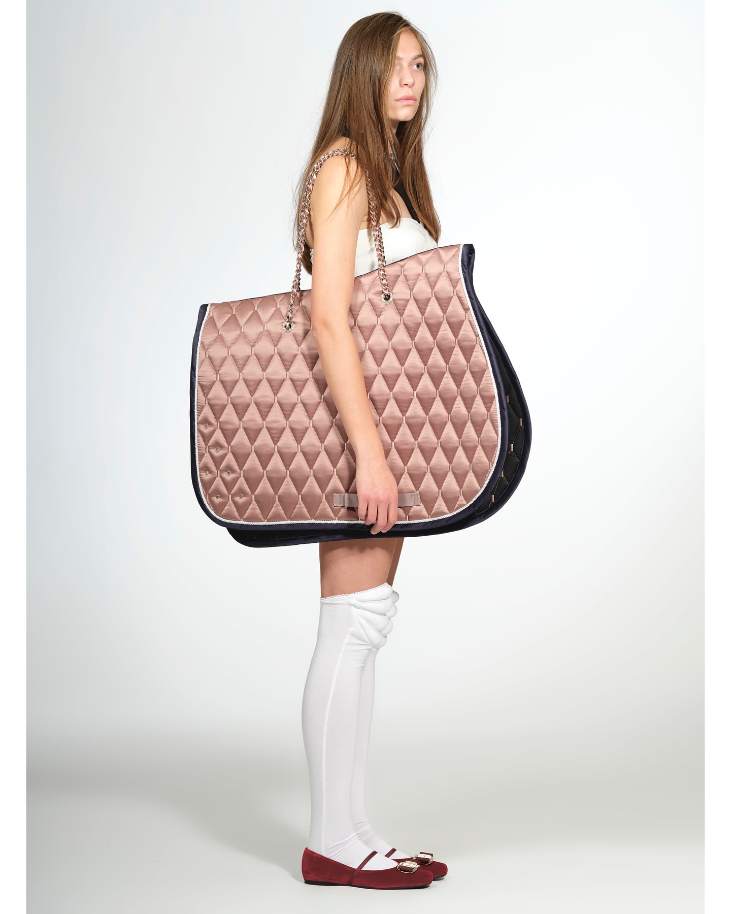 A young female model strikes a pose against a neutral backdrop, showcasing an oversized, quilted tote bag with chain straps.