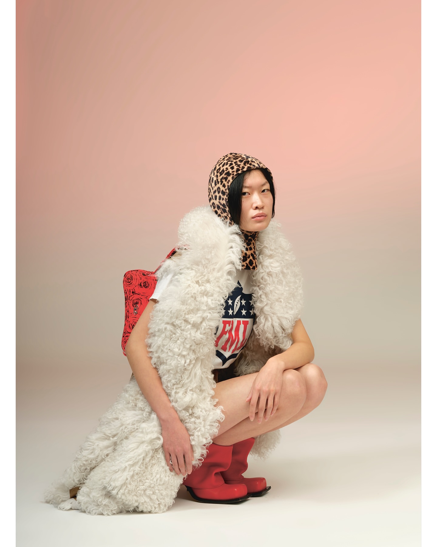 Portrait of a crouched Asian woman in a white t-shirt, red boots, and an animal print headscarf against a light gradient background.