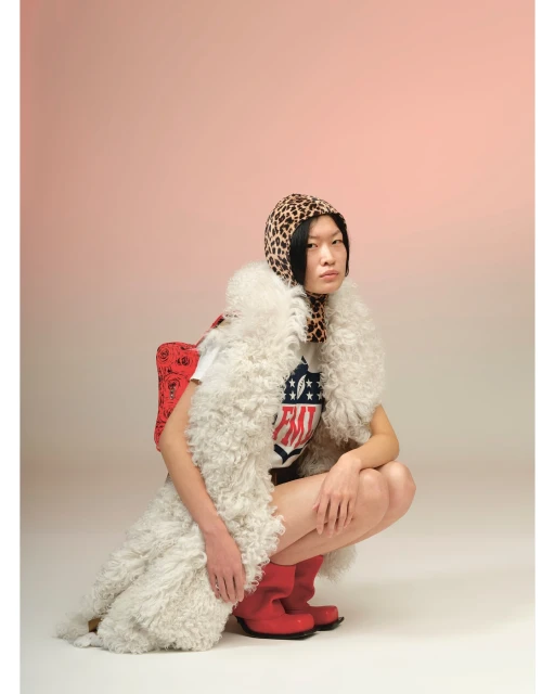 Portrait of a crouched Asian woman in a white t-shirt, red boots, and an animal print headscarf against a light gradient background.