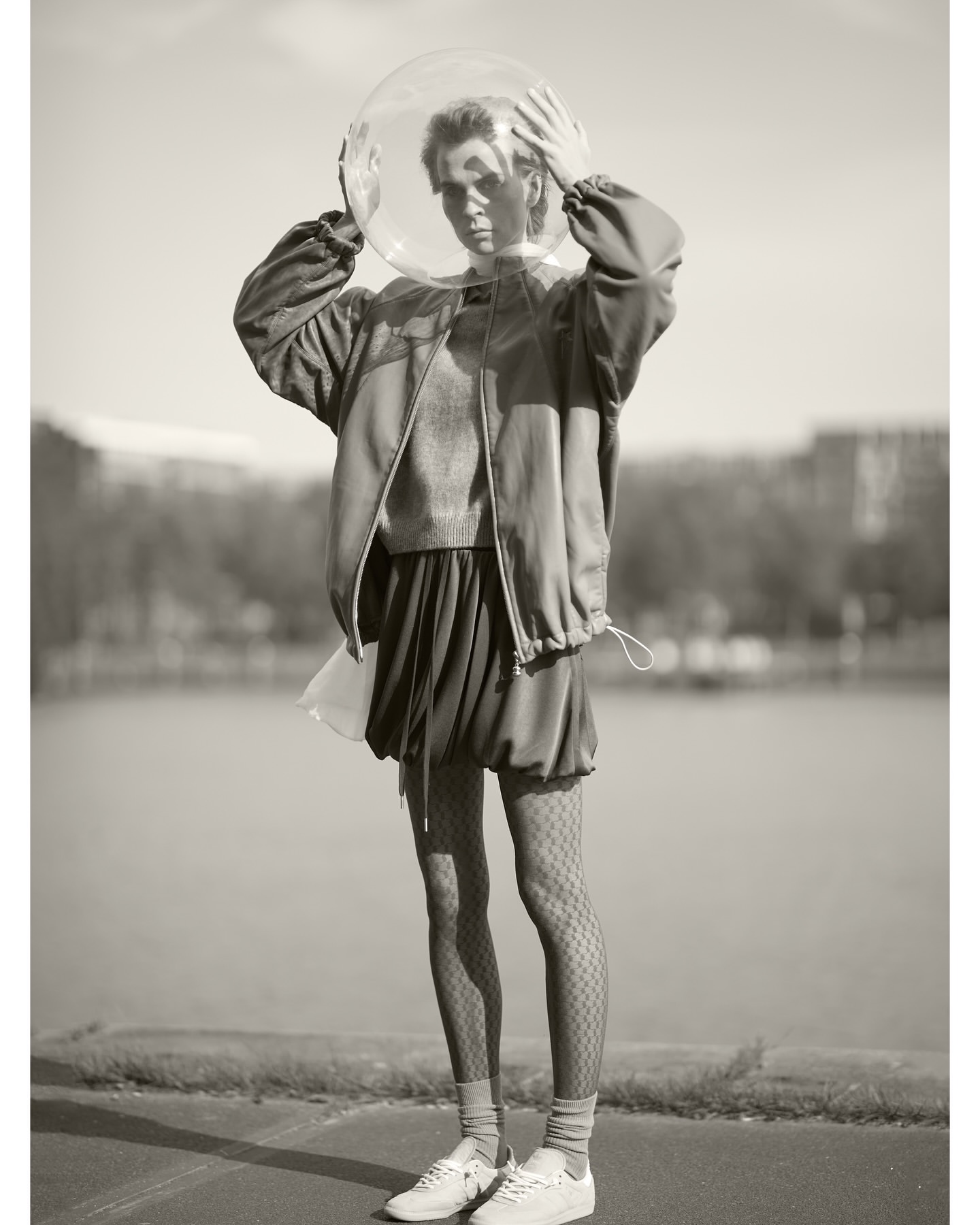 Monochrome portrait of a woman wearing a bomber jacket, skirt, sneakers, and a transparent sphere on her head in an urban park.