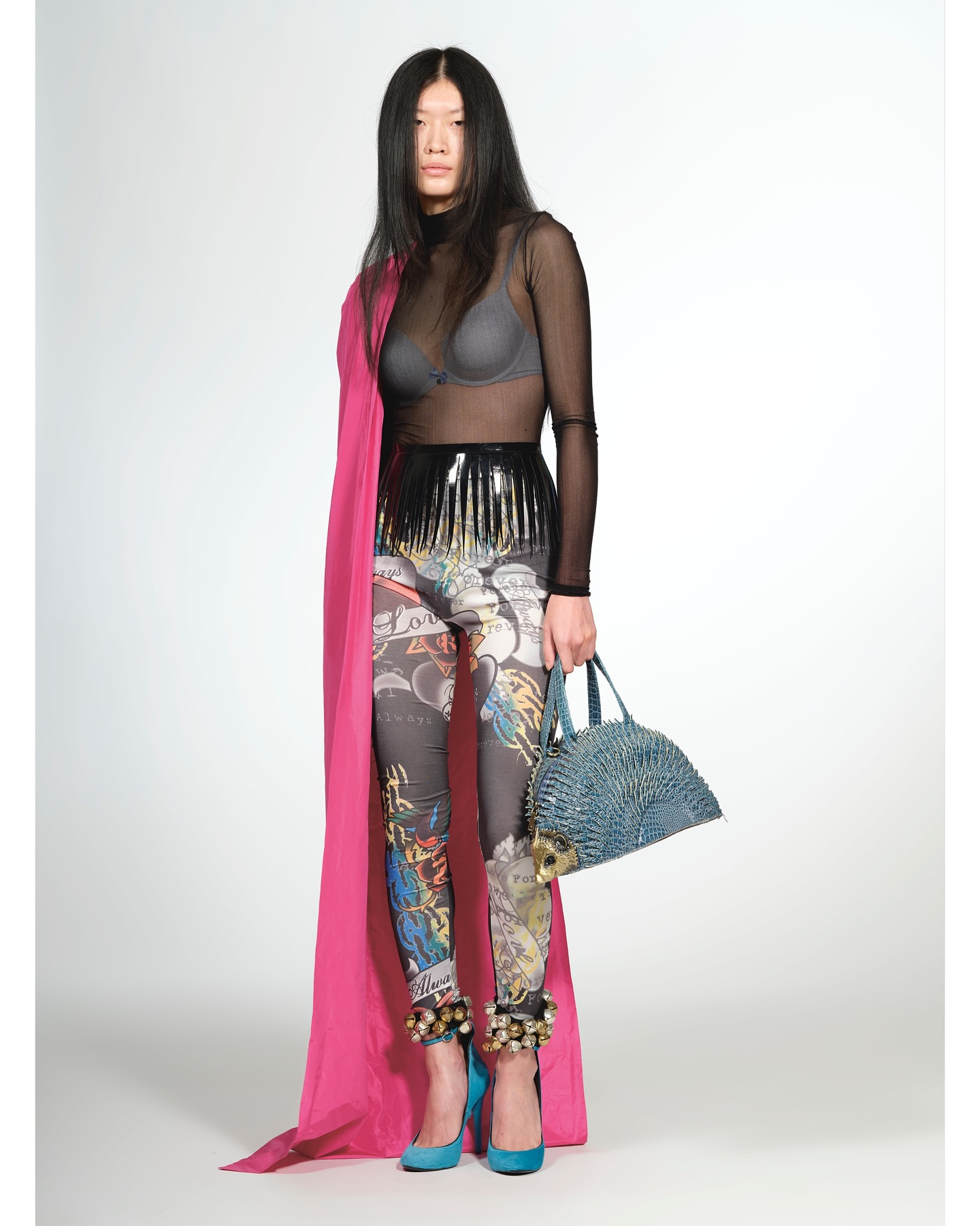 Full shot of an Asian female fashion model in a black top and patterned leggings, accessorized with a pink shawl, blue handbag and heels.