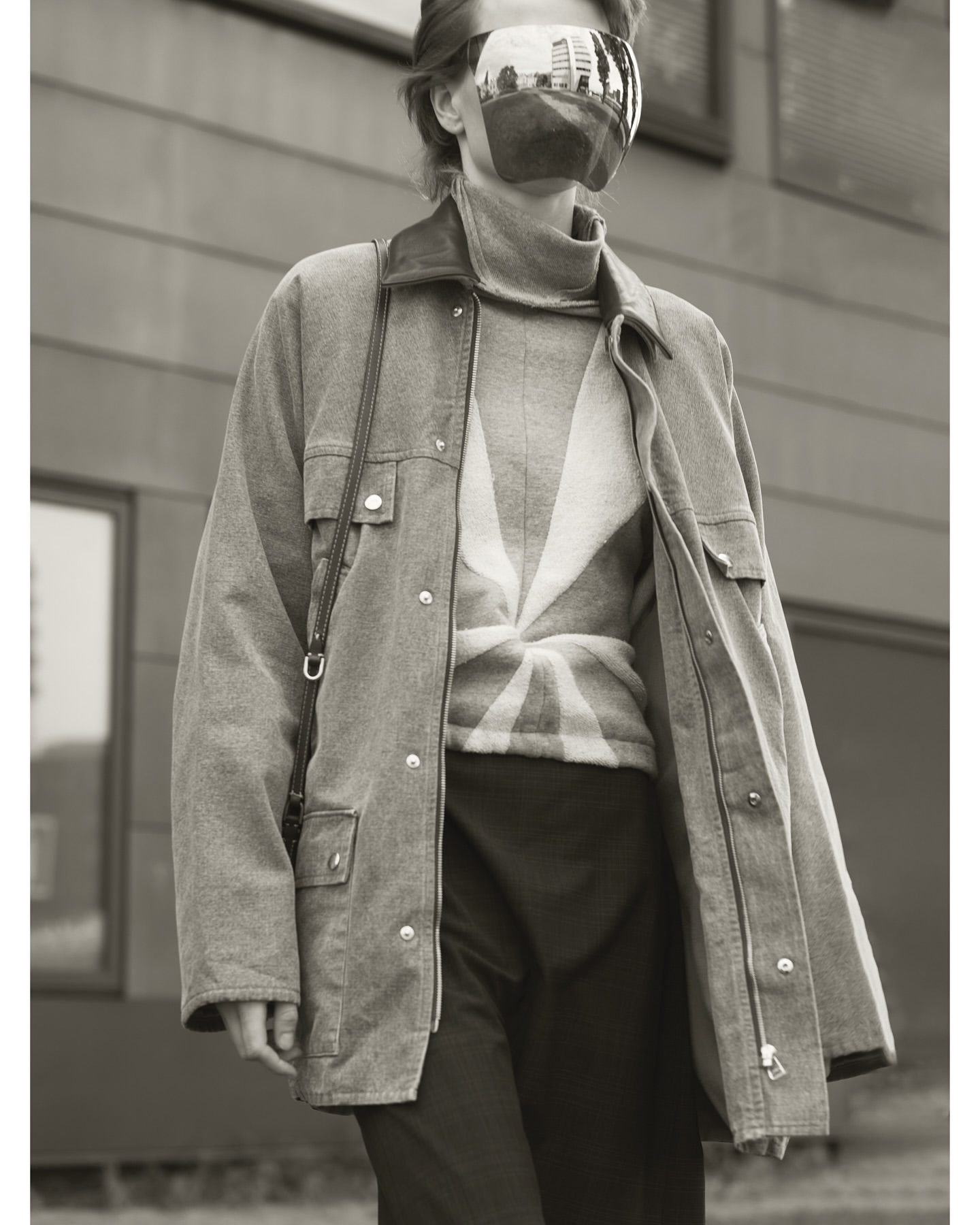 A person in a stylish outfit with a jacket and reflective visor stands outdoors, creating a fashion-forward and urban aesthetic.