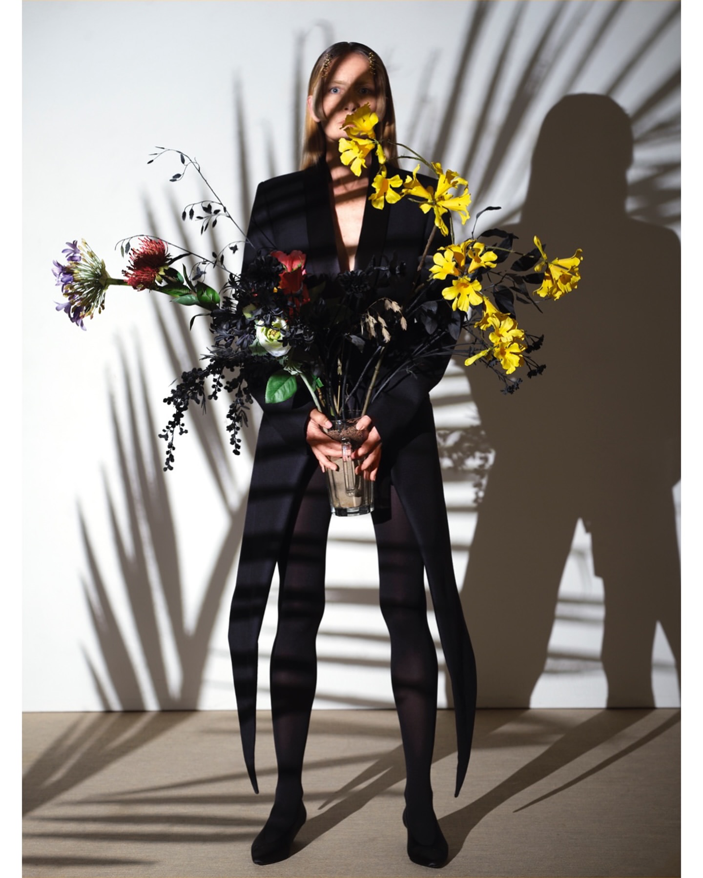 Woman in a black coat and tights holding a bouquet of colorful flowers in a vase, standing in front of a wall with palm leaf shadows.