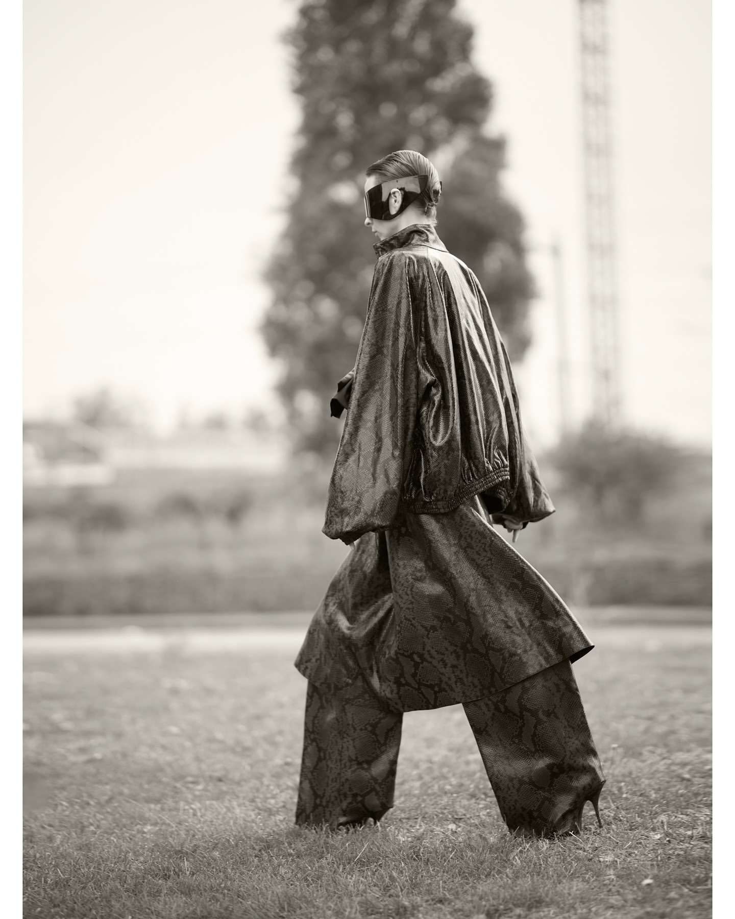 Monochrome image of a model wearing a snakeskin-patterned outfit and heels walking across a grassy field, wearing a black eye mask.