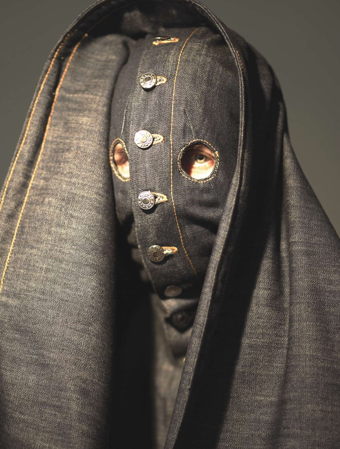 Person in full denim mask and hooded cloak with button accents, close-up studio shot. Fashion-forward and dramatic, the ensemble is striking.