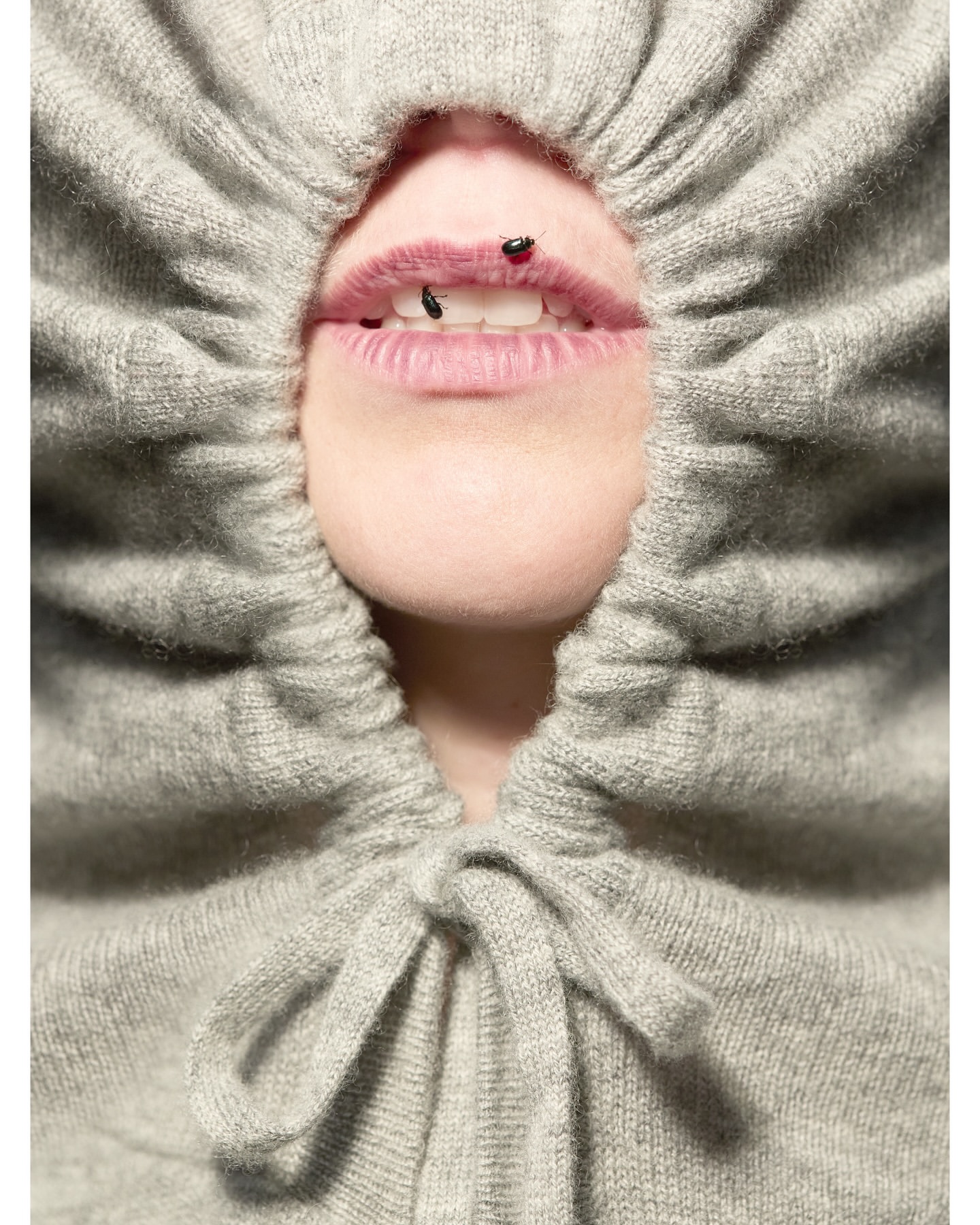 Close-up studio shot of a person's face with jewelry and a gray drawstring hood pulled around the face, emphasizing the mouth and facial features.