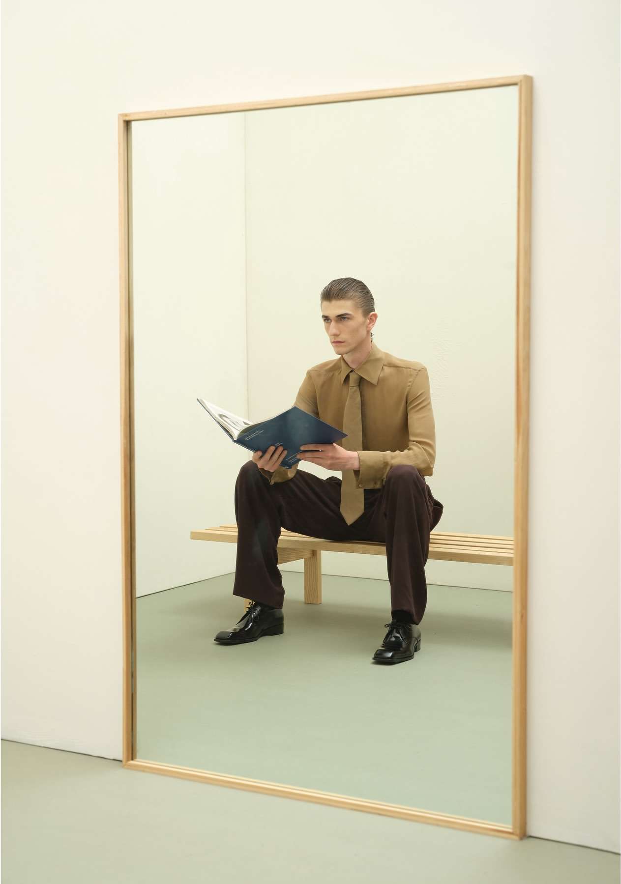 Young man in shirt and tie, seated on bench in front of large mirror, holding a book. Indoor setting, studio lighting, portrait orientation.