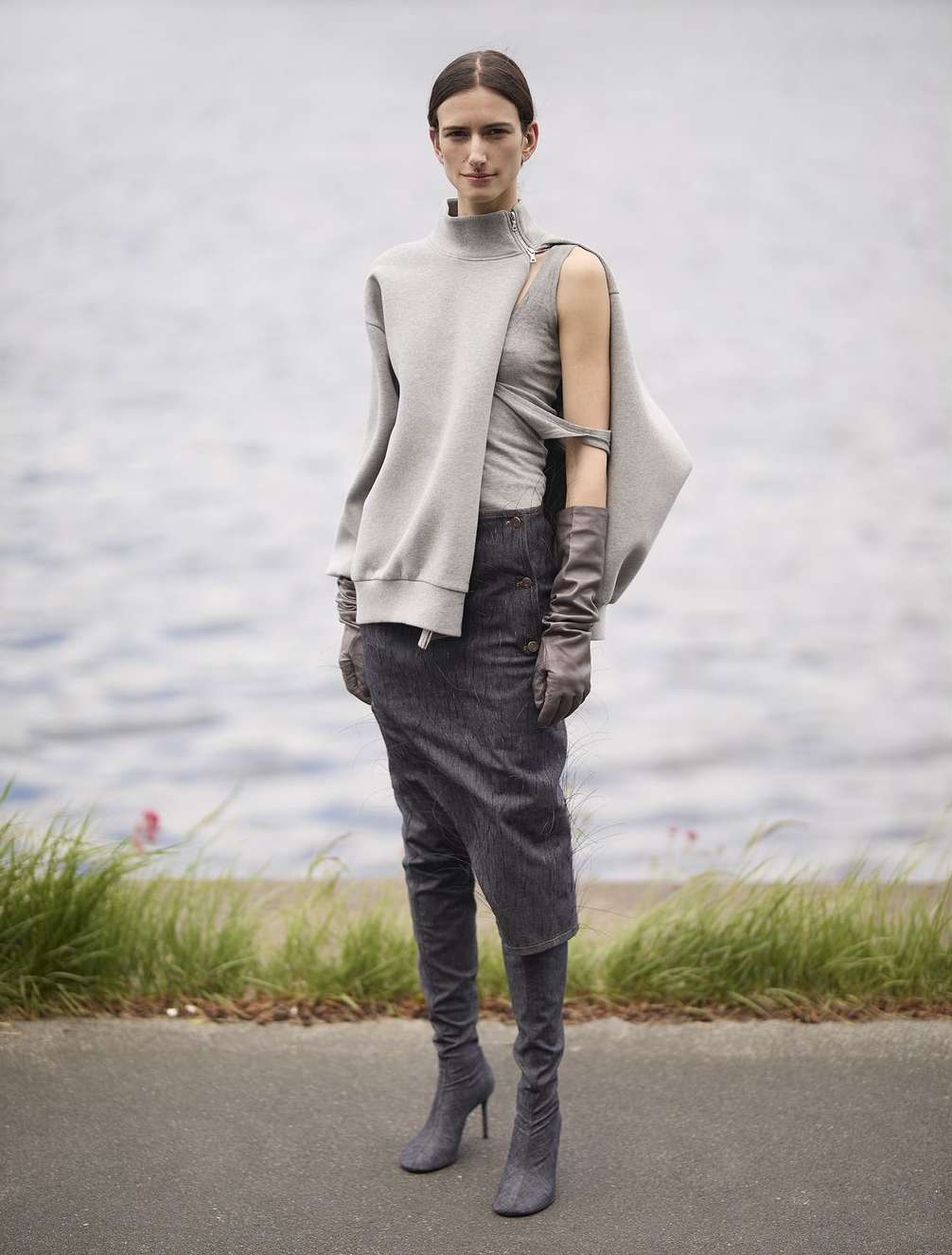 A stylish adult woman in a grey sweatshirt, skirt, gloves, and heeled boots poses outdoors with a calm expression and grey water background.