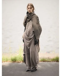 Model standing outdoors wearing layered taupe tunic trousers and a dark hooded cape in Paris by photographers Anuschka Blommers & Niels Schumm