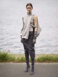 Model wearing a grey asymmetrical top, dark skirt, and grey boots standing by water, photographed by Anuschka Blommers and Niels Schumm.