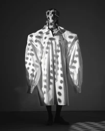 Monochrome portrait of a model in an oversized white dress with dramatic patterned shadows across the figure.