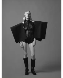 Model standing against a grey backdrop wearing a patterned black bodysuit with rigid structural black sleeves and dark boots.