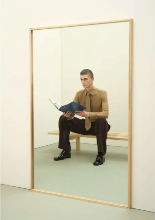 Man seated on a wooden bench holding an open magazine inside a large wood-framed mirror in a minimalist room.