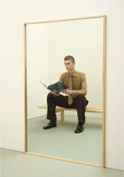 Man seated on a wooden bench holding an open magazine inside a large wood-framed mirror in a minimalist room.