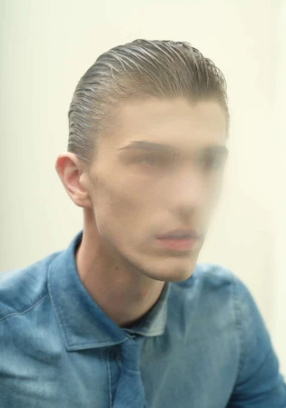 Close-up portrait of a young man with slicked-back dark hair wearing a blue shirt against a pale background.
