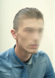 Close-up portrait of a young man with slicked-back dark hair wearing a blue shirt against a pale background.