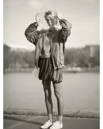 Model holding a transparent helmet over their head wearing a zip jacket and patterned tights outdoors.