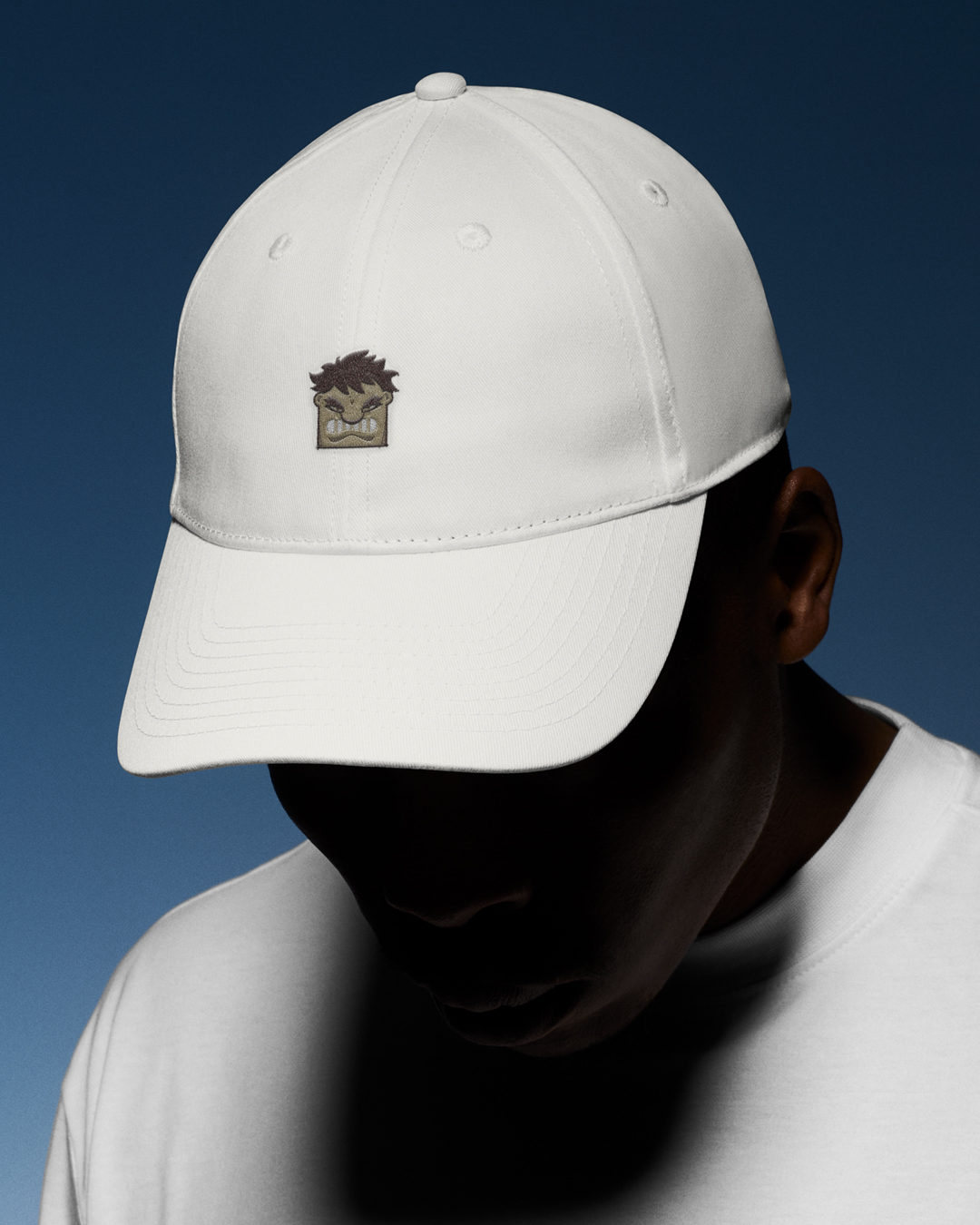 Man wearing a white baseball cap with a small embroidered cartoon graphic and a white t-shirt against a dark blue background